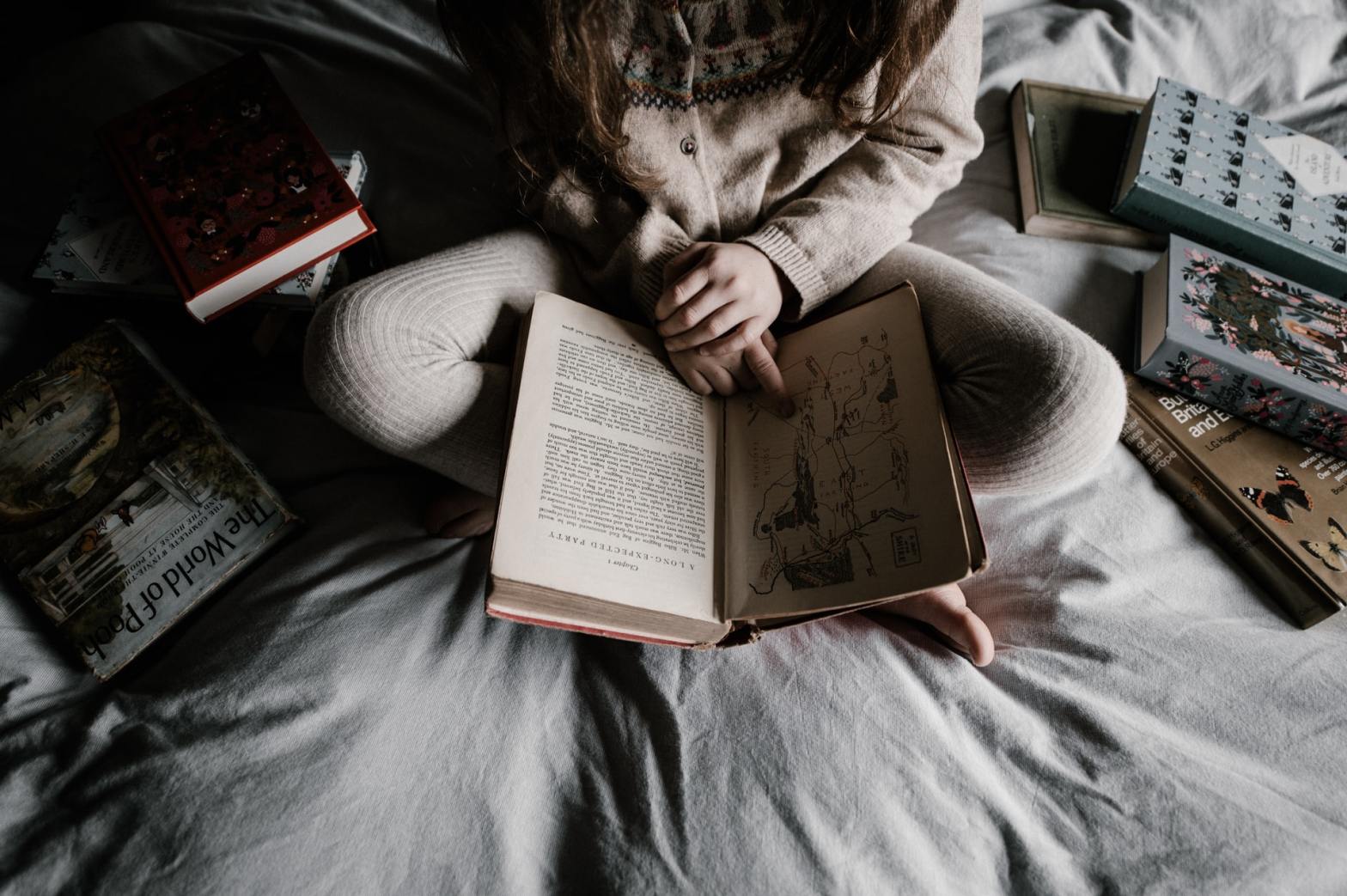 A girl sitting on a bed holding a book while being surrounded by other books